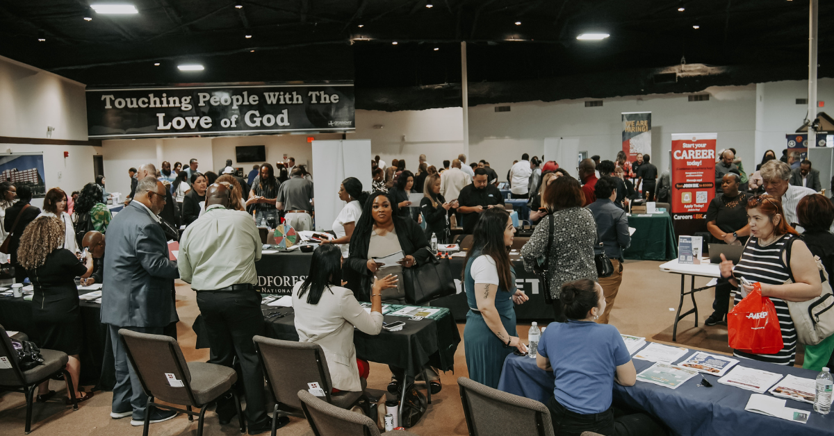 A busy job fair in a large indoor venue filled with diverse attendees visiting booths. A banner reading "Touching People With The Love of God" hangs above.