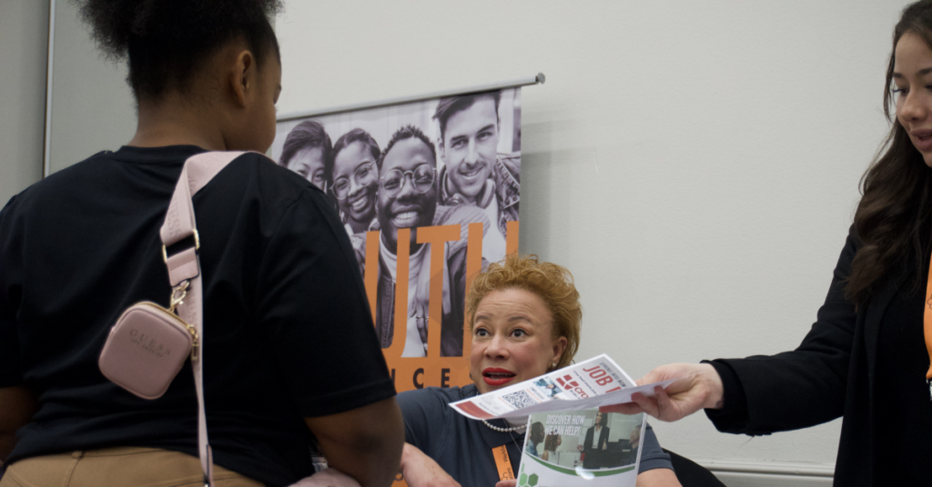 A woman talks to a visitor at a job fair booth, holding a flyer, with a banner showing smiling faces in the background.