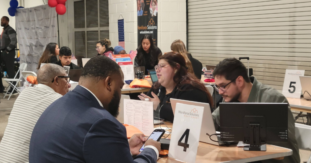People sit at tables in a job fair, engaging in discussions. Signs with numbers and "Workforce Solutions" are visible.