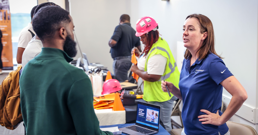 A woman in a blue polo shirt engages with a man at a job fair booth. Nearby, a person in a safety vest and helmet interacts at another table.