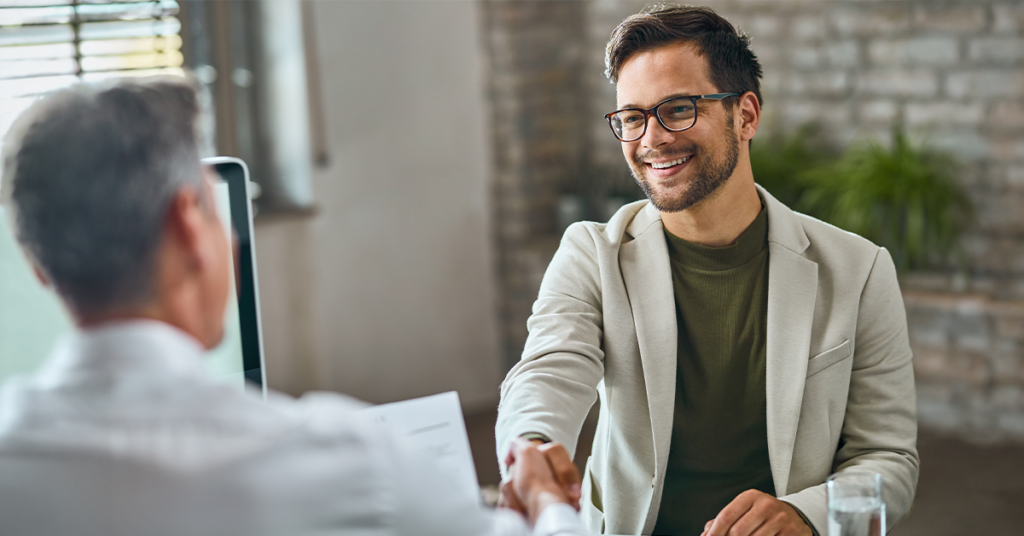 A man in glasses and a blazer smiles warmly while shaking hands with another person across a desk in an office setting.