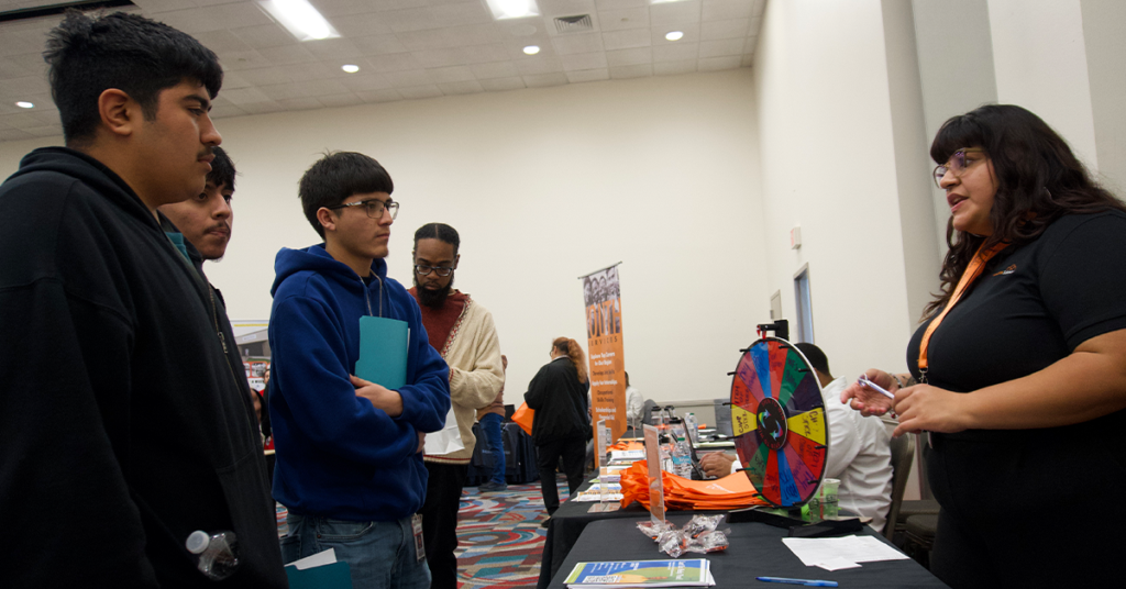 A group of young men listen attentively to a woman at a career fair booth. A colorful prize wheel is on the table, creating an engaging atmosphere.