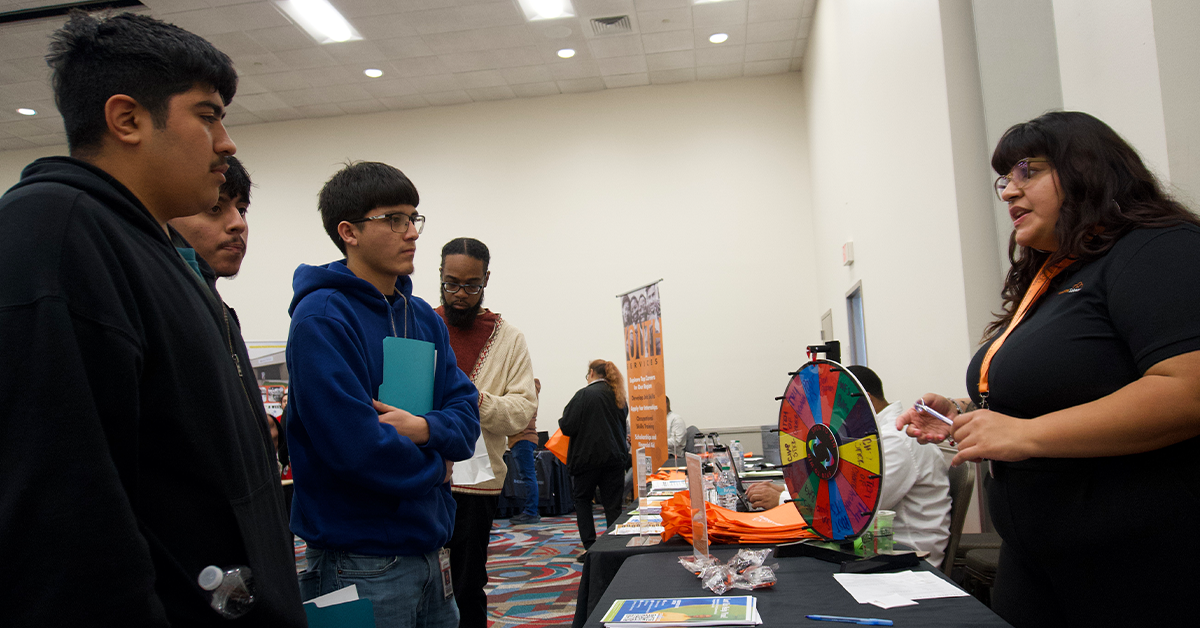 A group of young men listen attentively to a woman at a career fair booth. A colorful prize wheel is on the table, creating an engaging atmosphere.