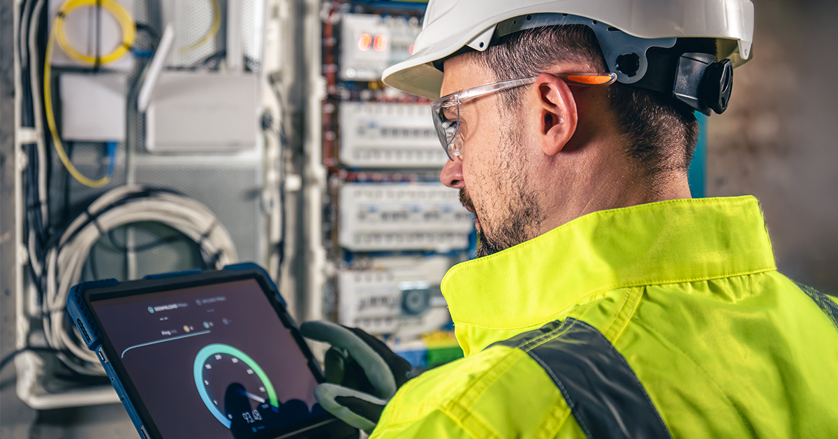 A worker in a high-vis jacket and hard hat uses a tablet in front of an open electrical panel, focusing on the screen with a digital gauge display, representing the Energy Industry.