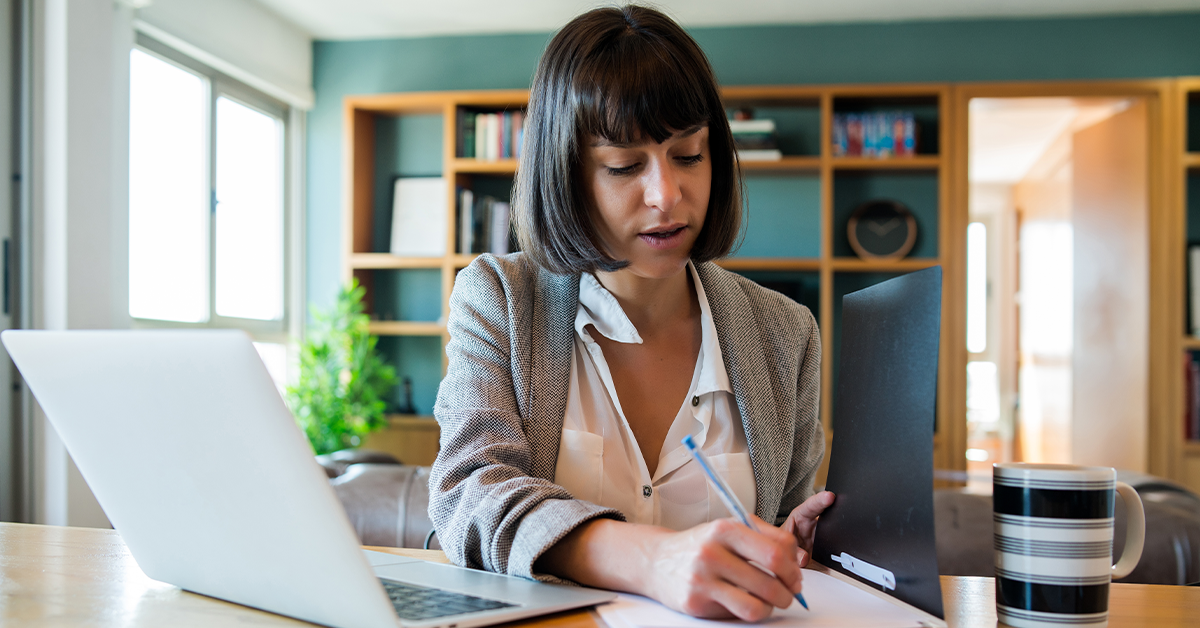 A woman with short brown hair works at a desk, writing notes on paper. A laptop and striped mug are beside her, with a bookshelf in the background.