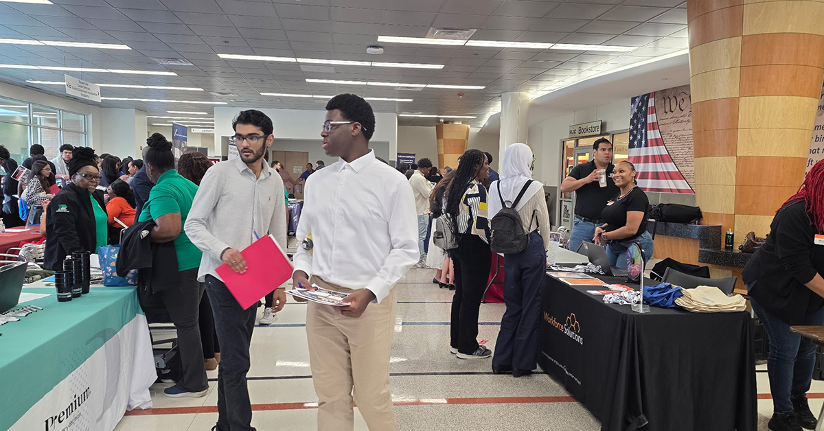 A bustling job fair with diverse attendees engaging at various booths. Two men in the foreground hold resumes, exuding professionalism and curiosity.