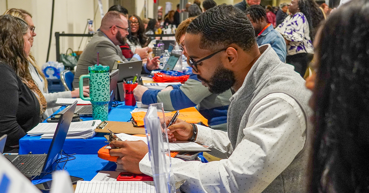 Skill Experience in Resume A group of people, seated at tables, engaged in discussions during a busy career fair. A man writes notes; laptops and colorful folders are visible.