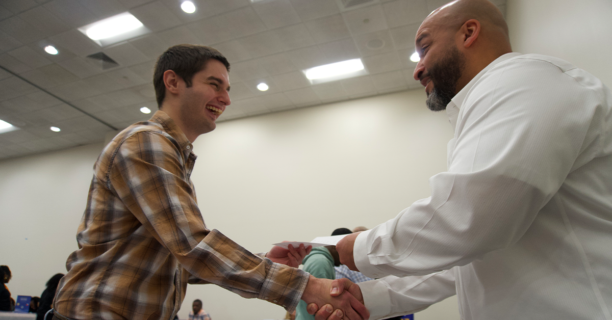 Two men smiling and shaking hands in a brightly lit room with a high ceiling.