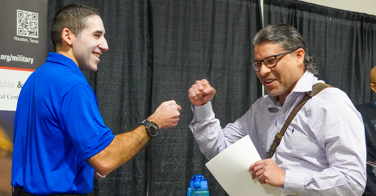 Work Values Two men happily engage in a fist bump at a professional event. One wears a blue polo shirt; the other a white shirt with brown strap and carries papers.