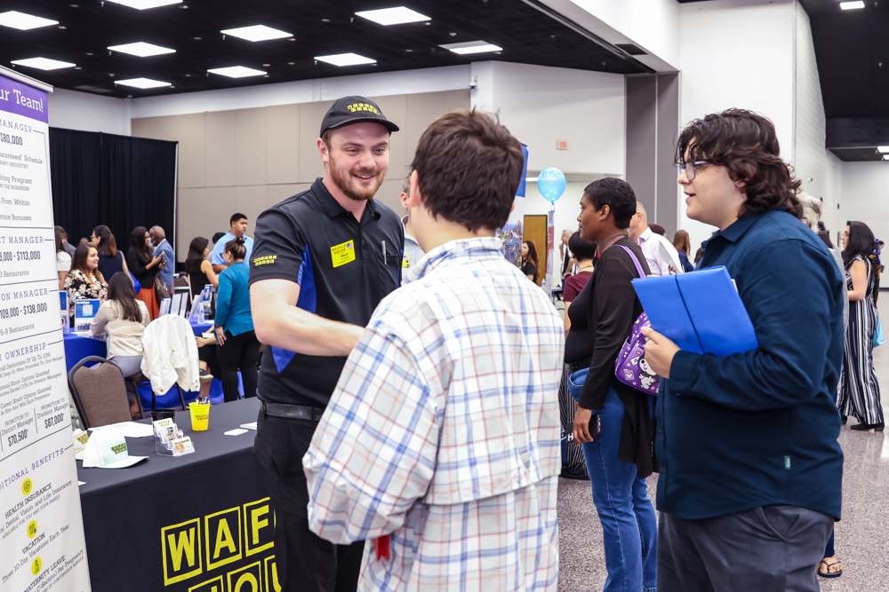A smiling man in a black and blue uniform shakes hands with an attendee at a job fair. The booth has company signs, and people converse in the busy background.