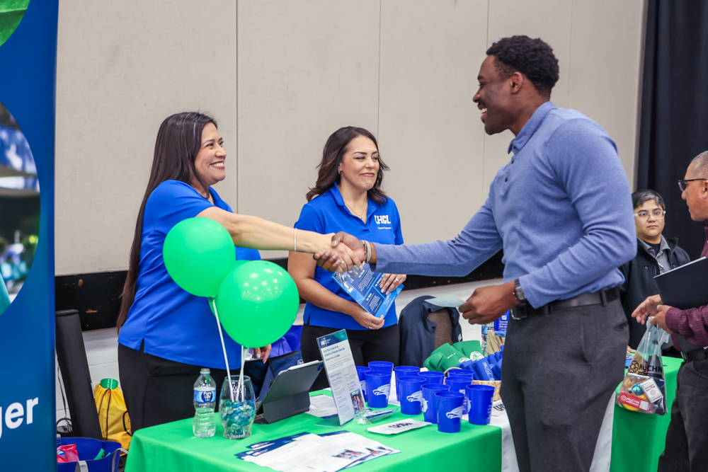 Man in a blue shirt smiling and shaking hands with a woman at a recruitment booth. Green balloons and promotional items are on the table.