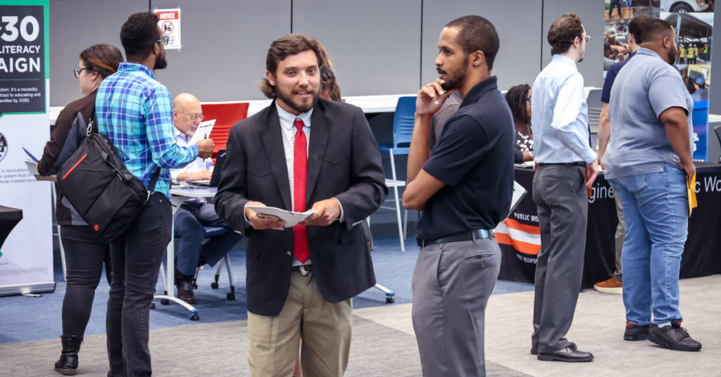 A busy job fair with professionals networking. Two men in conversation stand at the center, while others engage with company booths in the background.