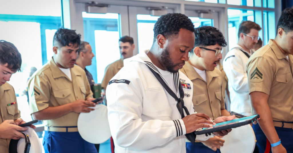 A group of military personnel in beige and blue uniforms, some holding hats, are gathered indoors. A man in a white naval uniform uses a tablet.