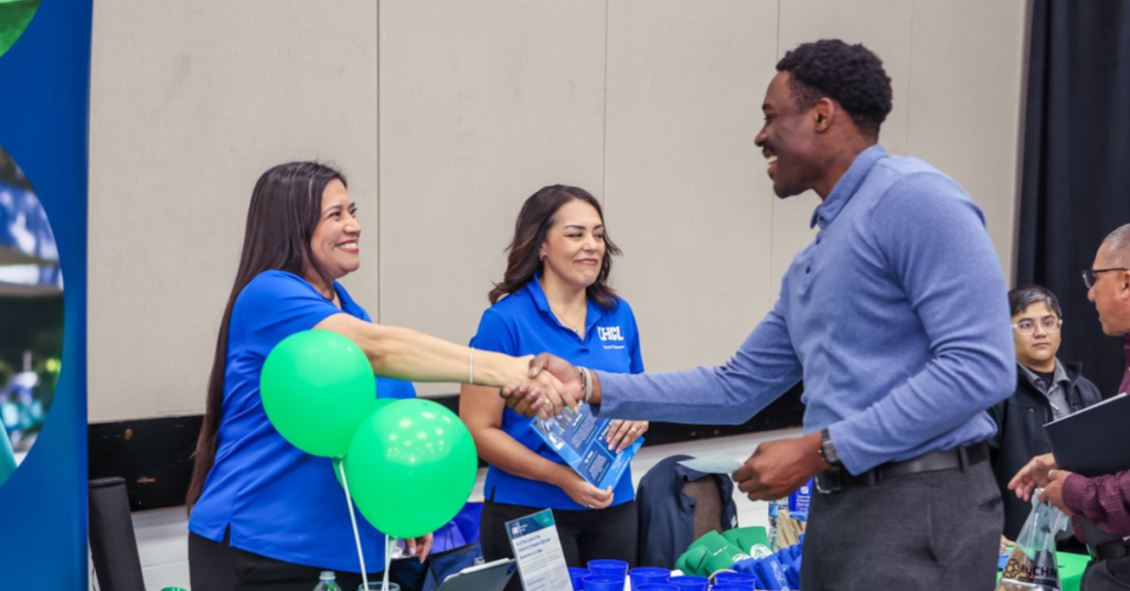 Man in a blue shirt smiling and shaking hands with a woman at a recruitment booth. Green balloons and promotional items are on the table.