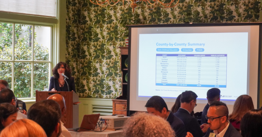 Juliet Stipeche speaks at a podium in a floral-wallpapered room, with a projector displaying a county summary chart. Attendees sit and listen attentively.