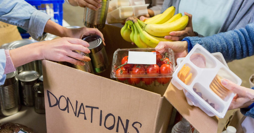 A group of hands places canned goods, bananas, grape tomatoes, and snacks into a cardboard box labeled "Donations."