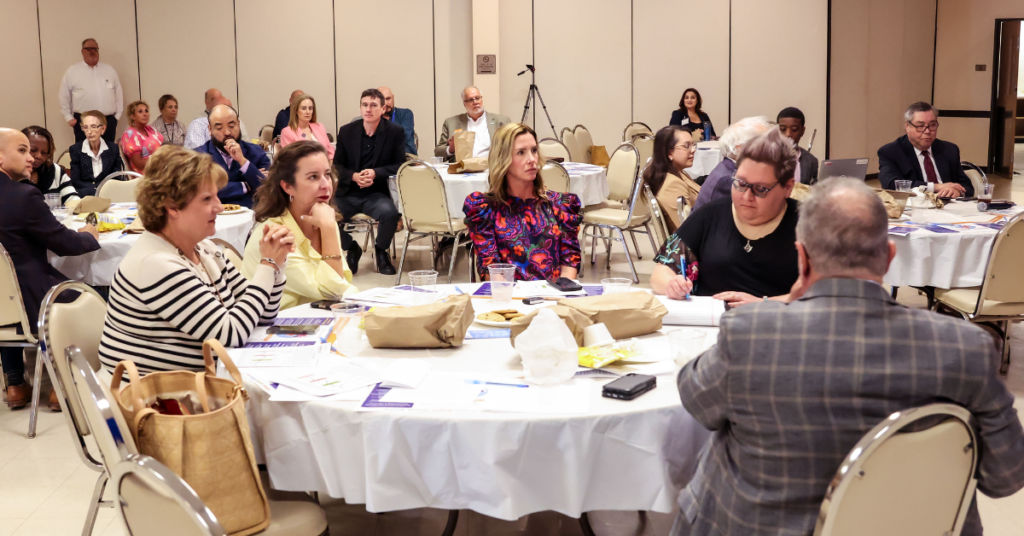 A group of people seated around tables in a conference room, attentively watching a presentation. Papers and snacks are on the tables, conveying focus.