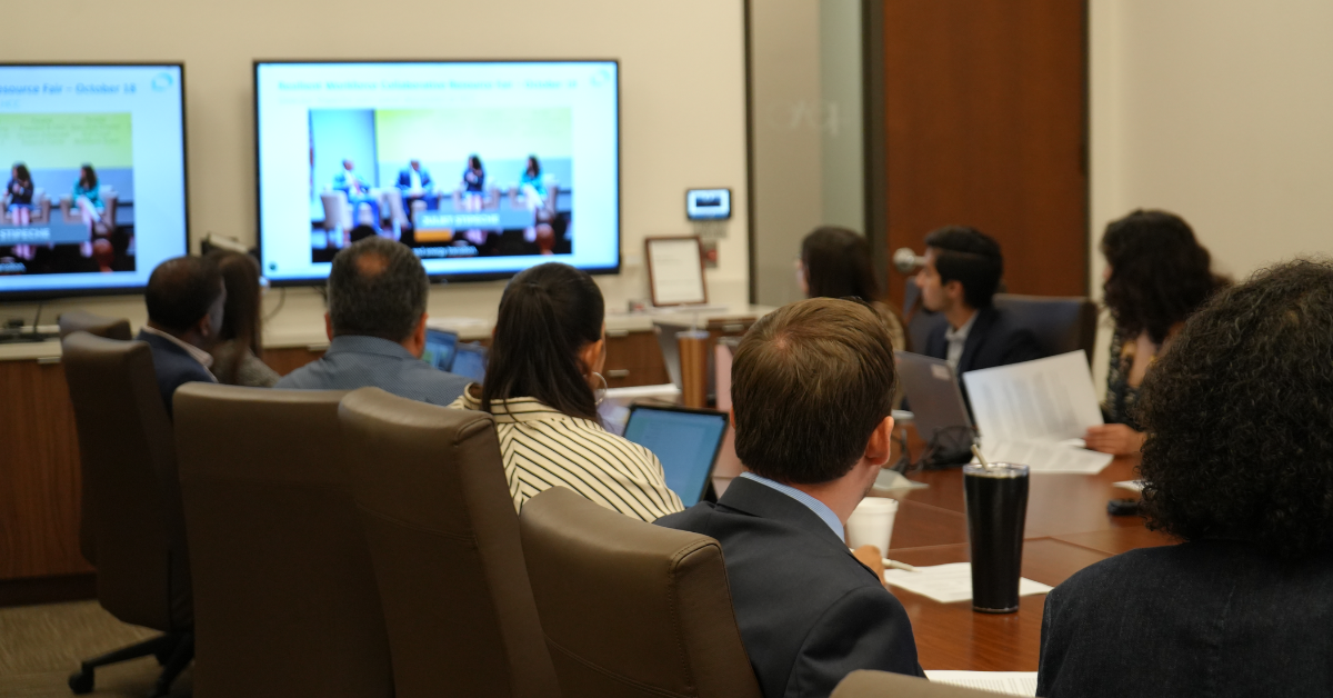 A group of people in a conference room face a large screen showing a virtual meeting. Attendees focus on the presentation.