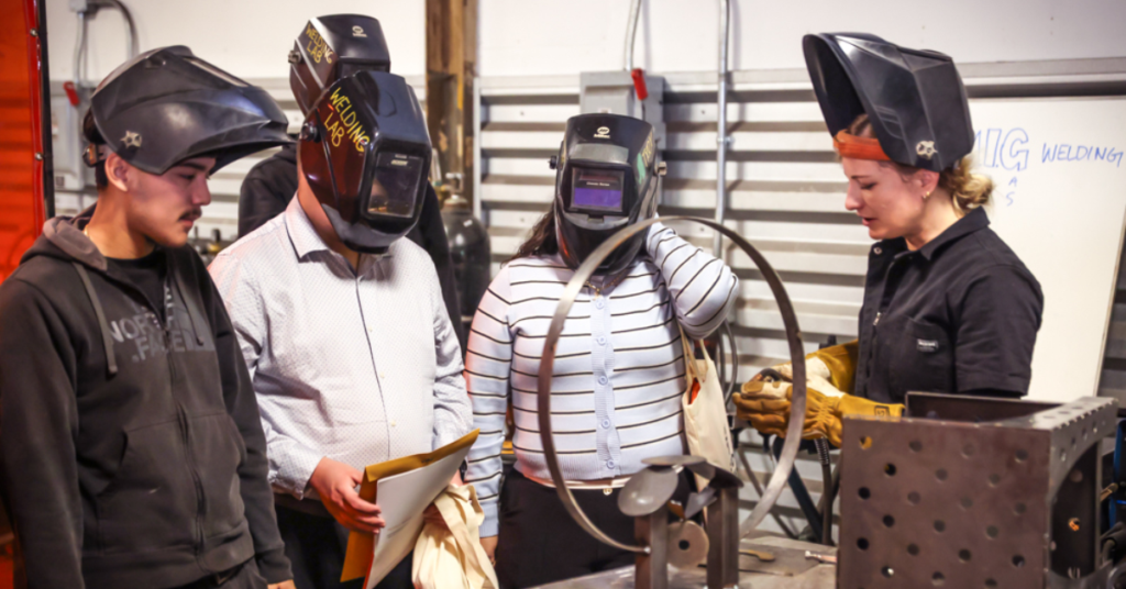A group of four people in welding helmets stands around a metal workshop table. One person instructs the others, creating a focused and educational atmosphere.