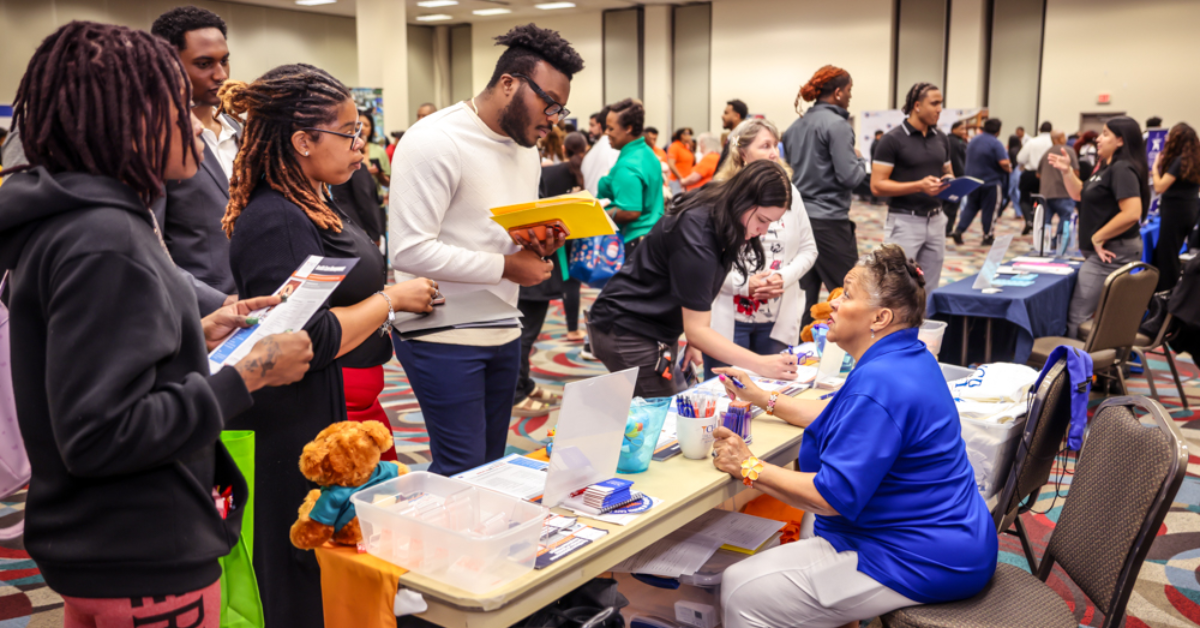 A bustling job fair scene with attendees engaging at various booths. People are interacting, holding pamphlets, creating a lively and hopeful atmosphere.