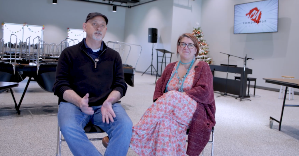 Carla Reus, Director of Empowerment for Compassion United, and Luke Reus, CEO of Compassion United, sit in a modern room with stacked chairs and a Christmas tree. They appear to be conversing.