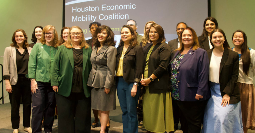 Group photo of attendees and regional leaders at the Houston Economic Mobility Summit. Juliet Stipeche stands at the center alongside workforce and community partners gathered in front of a presentation screen displaying “Houston Economic Mobility Coalition.”