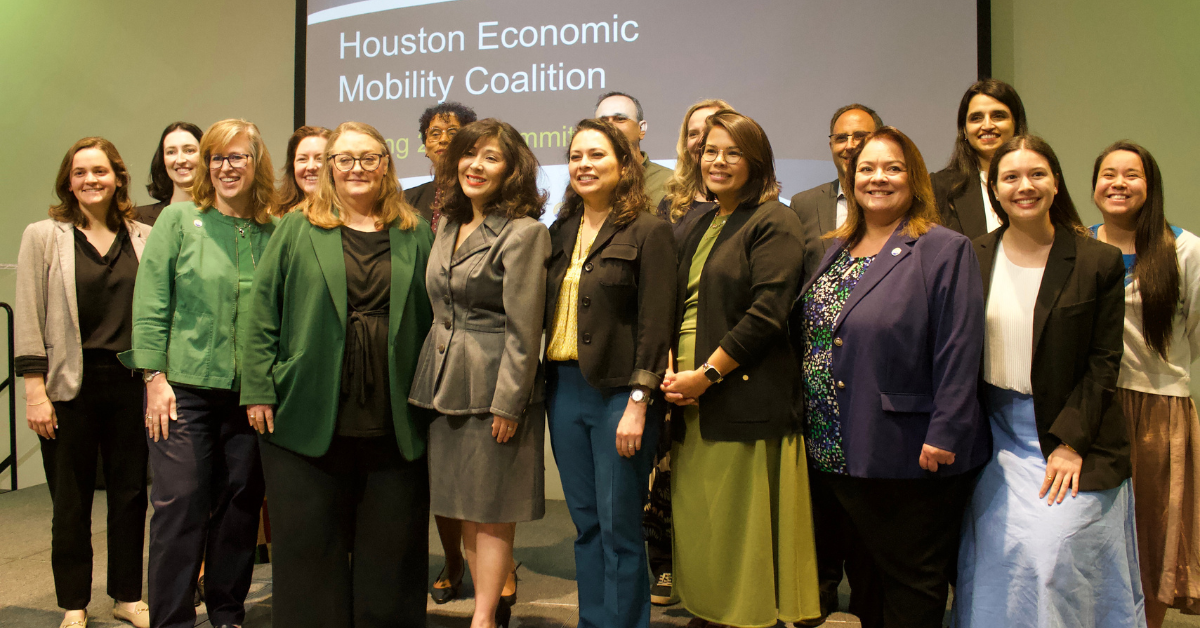 Group photo of attendees and regional leaders at the Houston Economic Mobility Summit. Juliet Stipeche stands at the center alongside workforce and community partners gathered in front of a presentation screen displaying “Houston Economic Mobility Coalition.”