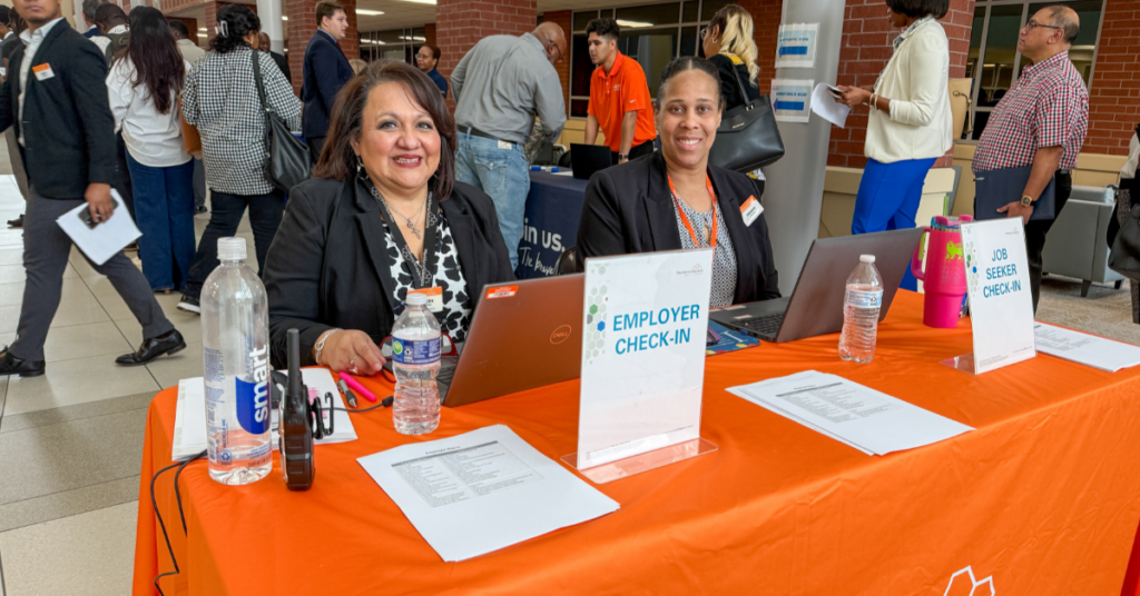 Two women seated at a check-in table with laptops, smiling at a job fair. Signs for "Employer Check-In" and "Job Seeker Check-In" are visible.