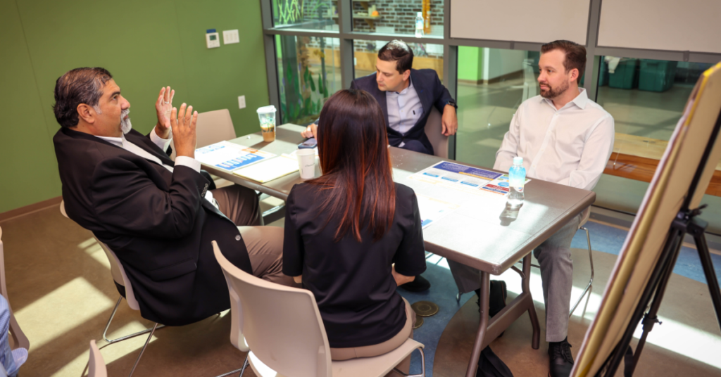 Four people in business attire are in a meeting room with a green wall. One man gestures expressively while others listen attentively at a table.