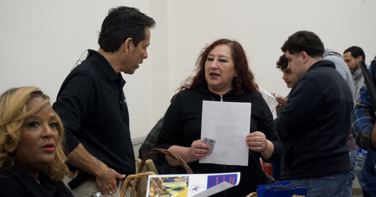 In a busy room, two people engage in conversation, one holding a paper. Others are in the background. The atmosphere is focused and lively.