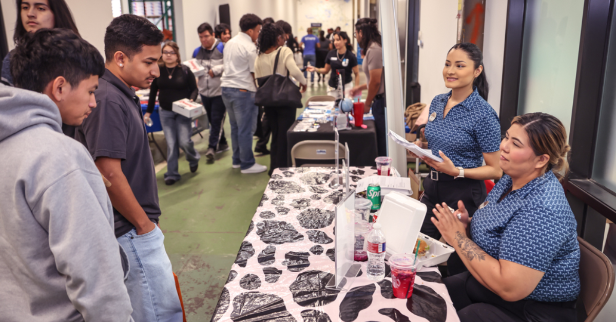 Youth Job Fair A bustling job fair with two women at a table interacting with attendees. People line up, some holding resumes, in a lively, engaging atmosphere.