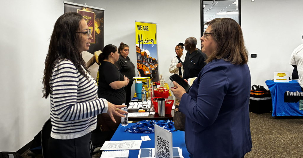 Two women converse at a job fair booth with "We Are Hiring" signs. Others engage nearby, creating a bustling, professional atmosphere.