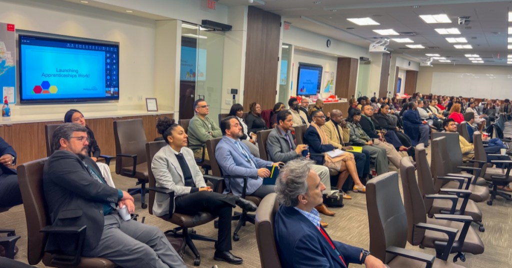 A diverse group of people seated in a well-lit conference room, attentively listening to a presentation titled "Launching Apprenticeships Week." The atmosphere is focused and professional.