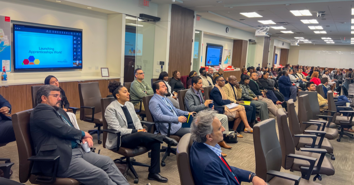 Apprenticeship A diverse group of people seated in a well-lit conference room, attentively listening to a presentation titled "Launching Apprenticeships Week." The atmosphere is focused and professional.