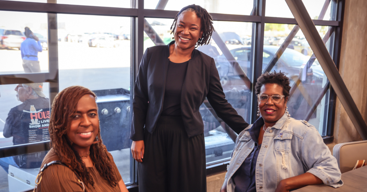Three women smiling warmly, gathered indoors by large windows. One stands in business attire, the others sit casually.