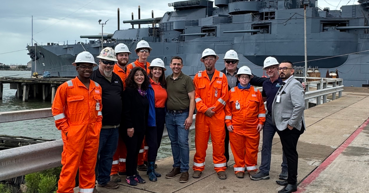 Juliet Stipeche, Executive Director of the Gulf Coast Workforce Board and Workforce Solutions Gulf Coast, standing next to workers in orange coveralls and hard hats. They pose happily in front of a large gray battleship docked by the water.