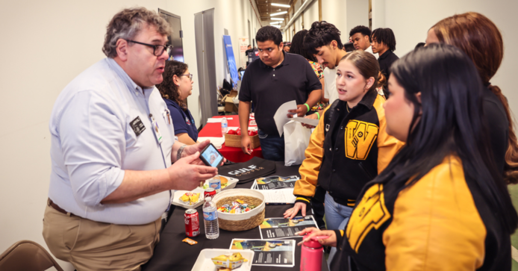 A man at a career fair booth talks to three young individuals. The table has brochures, candy, soda, and a basket.