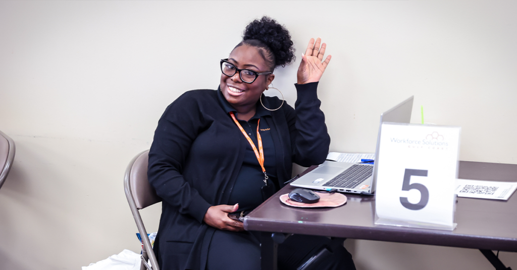 Smiling woman in glasses and black outfit sits at a desk with a laptop, raising her hand.