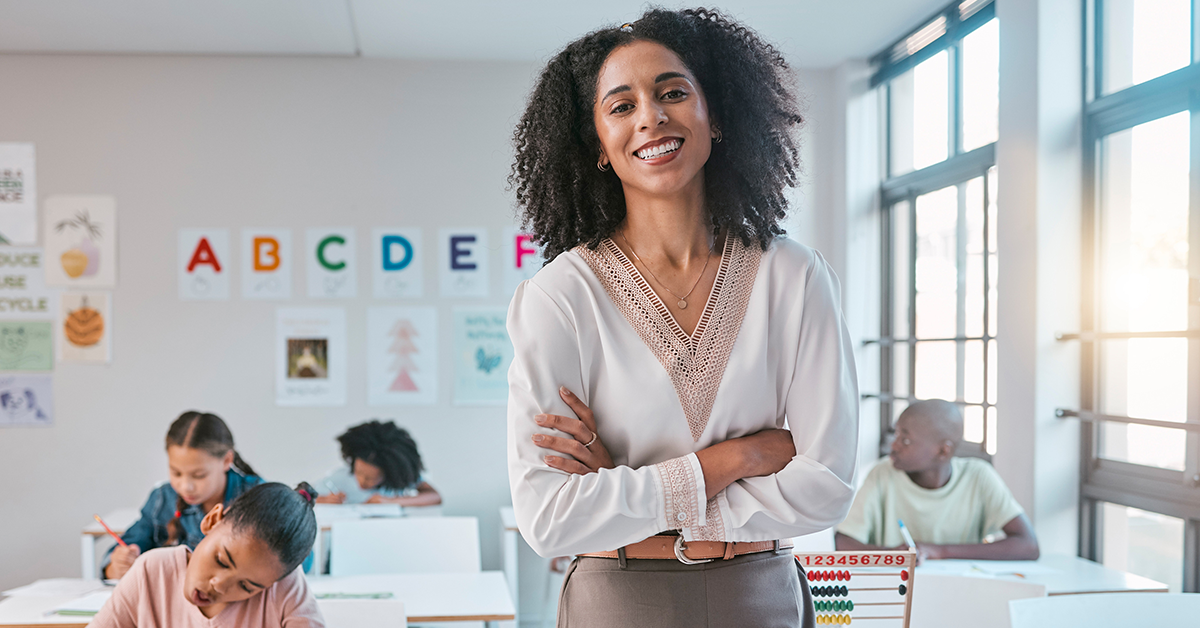 Education Industry. Smiling teacher stands confidently in a bright classroom with arms crossed. Children sit at desks working. Walls display colorful letters and posters.