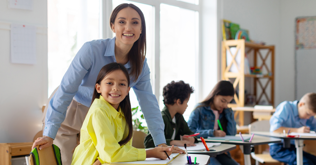 A smiling teacher leans over a young girl in a classroom. The girl, in a yellow shirt, smiles while classmates focus on their work in the background.