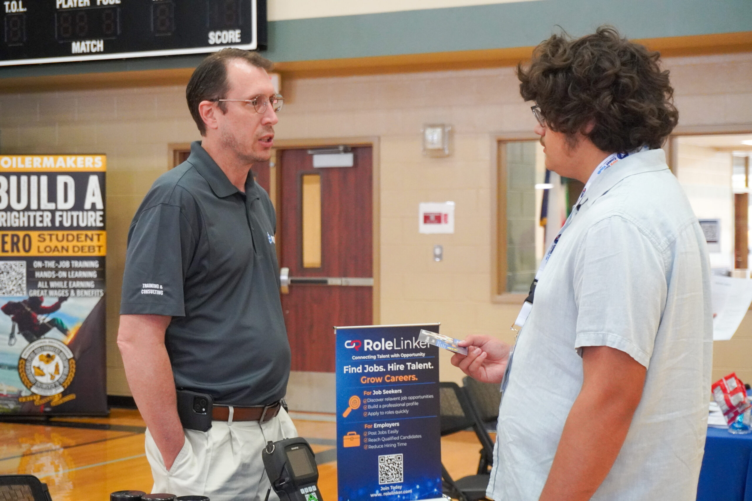Two men engaged in conversation at a job fair. One wears a dark polo and the other a light shirt with a lanyard. Banners about career opportunities are in the background.