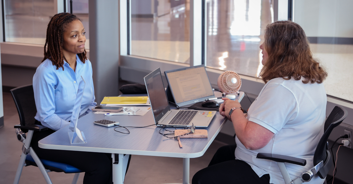 Interview Two people sit across from each other at a small desk, engaged in conversation. Laptops and documents are on the table in a modern office setting.