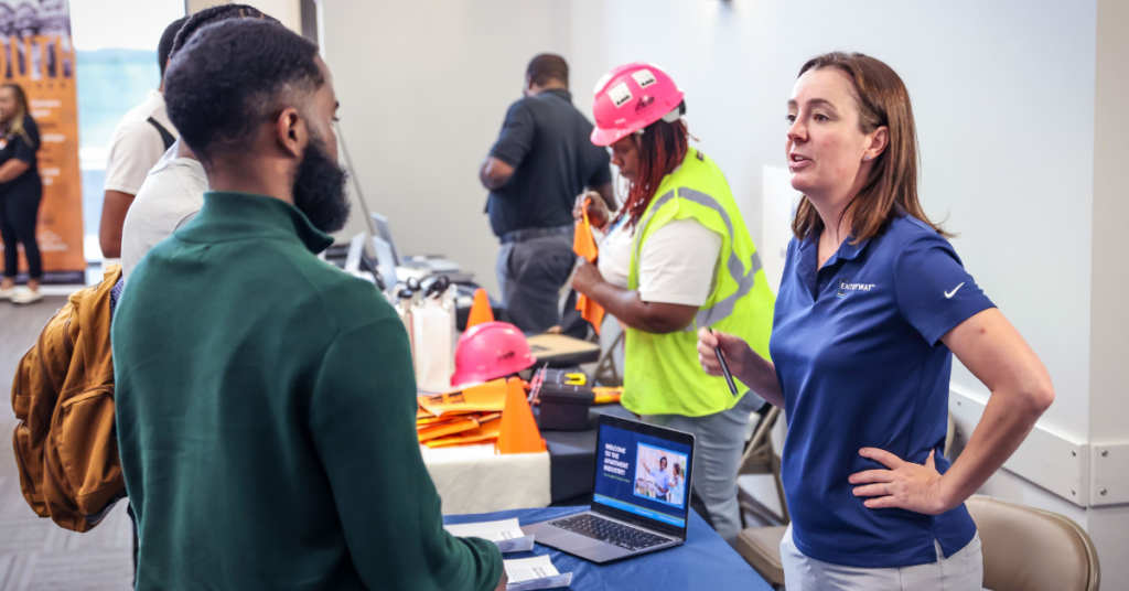 A woman in a blue polo talks to a man at a career fair booth. Nearby, a worker in a pink hard hat interacts with materials. The setting is lively and engaging.