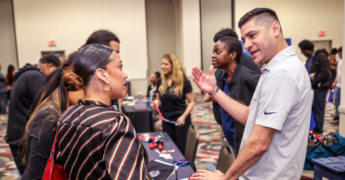 Job Fair A man and woman engage in conversation at a busy networking event, with people mingling in the background.