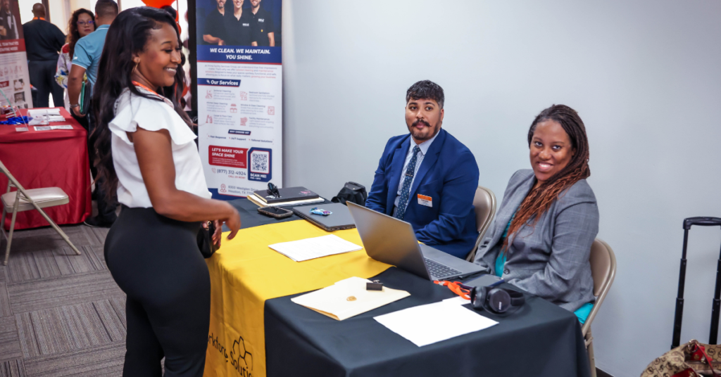 A woman in business attire stands smiling at a table with two seated people behind a laptop. The setting is a professional conference or career fair.