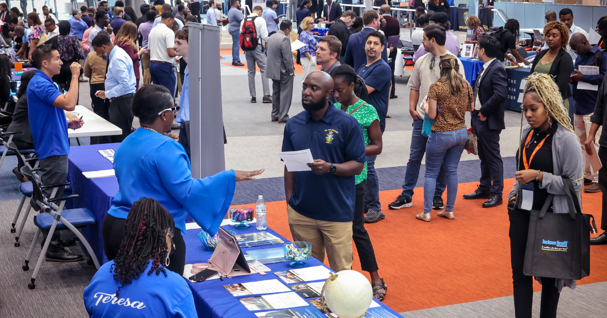 A busy career fair scene with people interacting at various booths. A woman in blue on the left speaks to a man holding a paper.