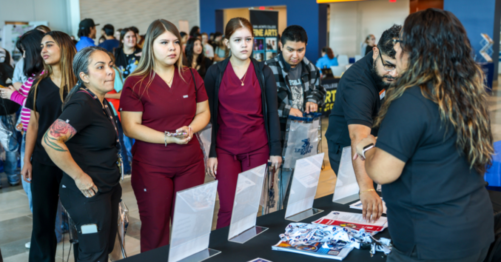 A group of people stand at a table at a Hiring Event. Two young women in maroon outfits look intently at brochures while others engage with staff across the table.