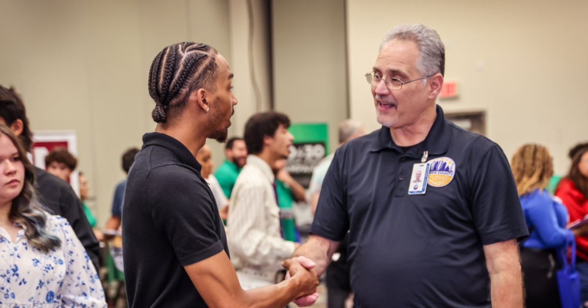 Training Grant Two men are shaking hands at a busy indoor event. Both wear dark polo shirts. The scene conveys a friendly, professional interaction.