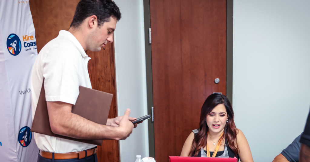 A job seeker stands holding a folder and phone, speaking to a career specialist who is seated and working on a laptop.
