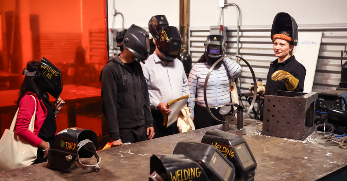 A group of five people wearing welding helmets stand around a metal workshop table, listening to an instructor.